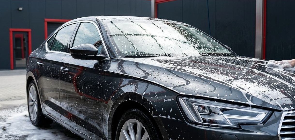 A glossy dark sedan covered in thick white foam inside a modern car wash bay.