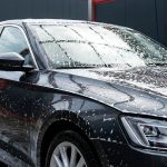 A glossy dark sedan covered in thick white foam inside a modern car wash bay.