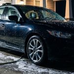 A dark sedan being washed with soap and water in a brightly lit driveway at night.
