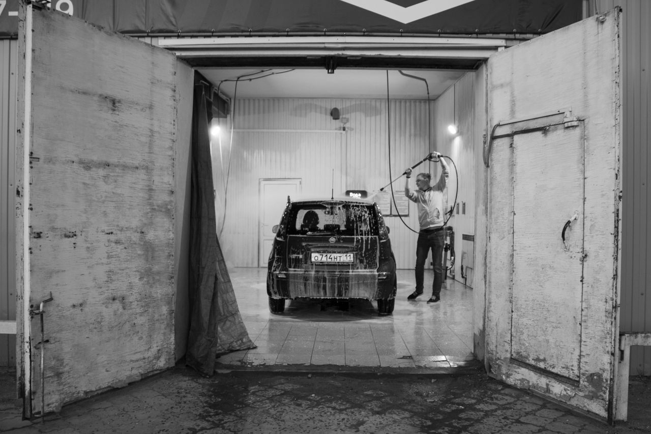 Black and white image of a man washing a car in a garage setting.
