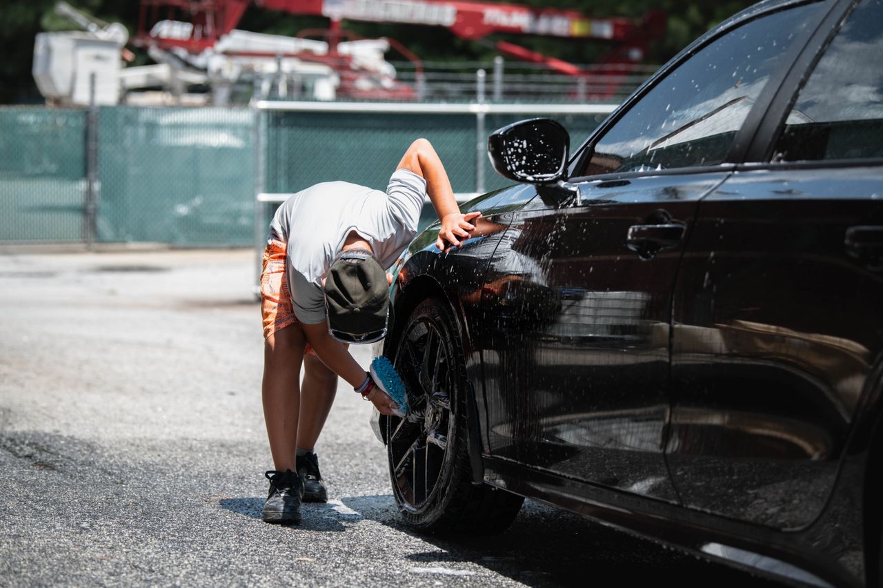 A person cleans a black car's wheels during a sunny day outdoors.