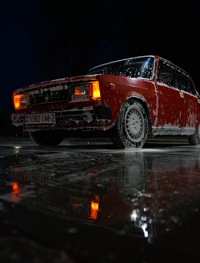 Vintage red car being washed at night, reflecting on wet pavement with lights on.