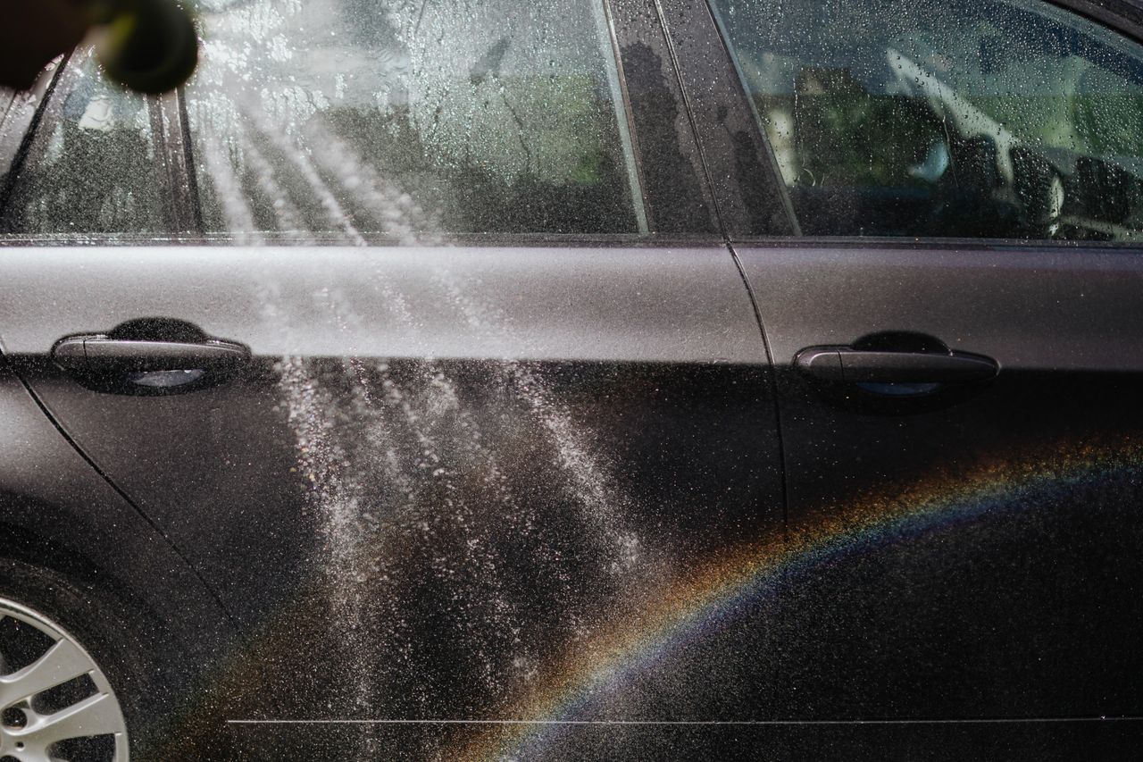 Close-up of a car being washed with a visible rainbow in sunlight.