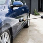 A wash gun sprays water across a glossy dark blue car in a driveway.