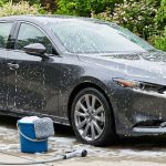 A dark car covered in foam beside a garden, bucket, and water-saving nozzle.