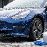 Foam-covered blue car being washed beside a bucket, mitt, and hose.