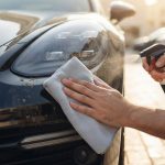 A dark car bumper being cleaned with a towel and spray bottle in sunlight.