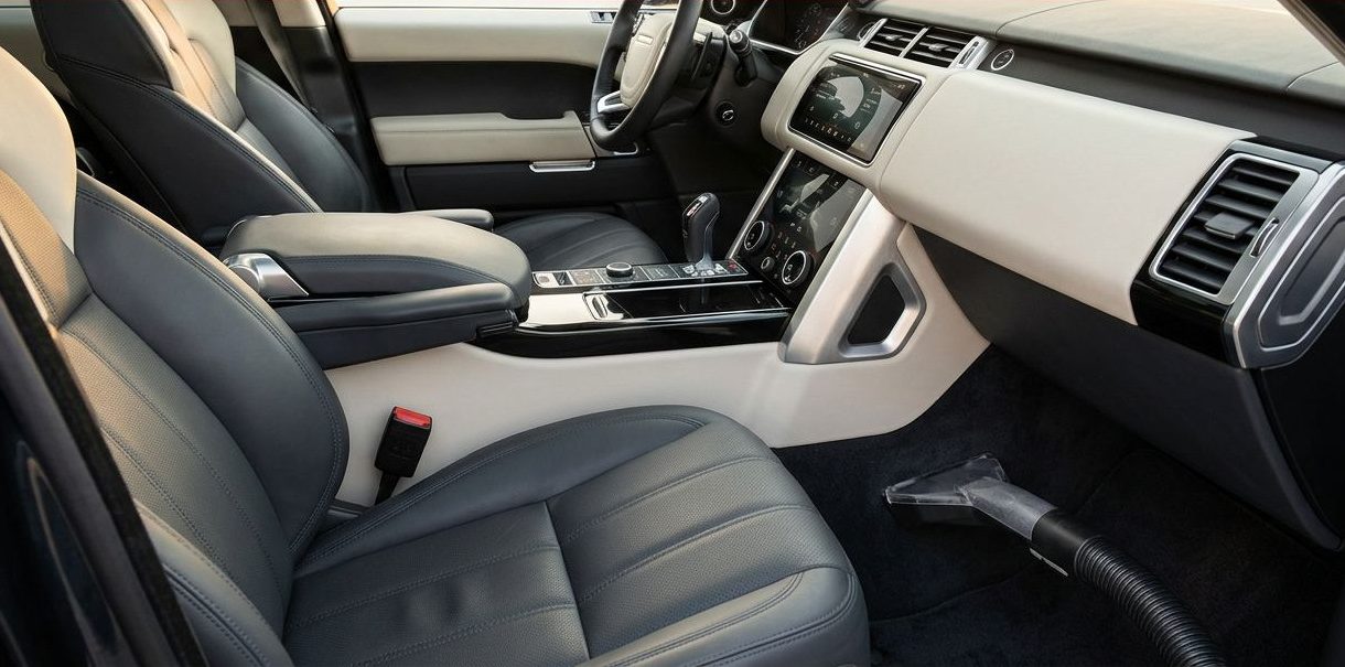 Vacuum nozzle cleaning a spotless car carpet beside leather seats and a visible dashboard touchscreen.