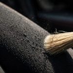 A red-handled brush lifts the fibers on a charcoal car seat.