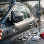 A hand photographs a scratched side mirror beside printed photos and a receipt on a wet car hood.
